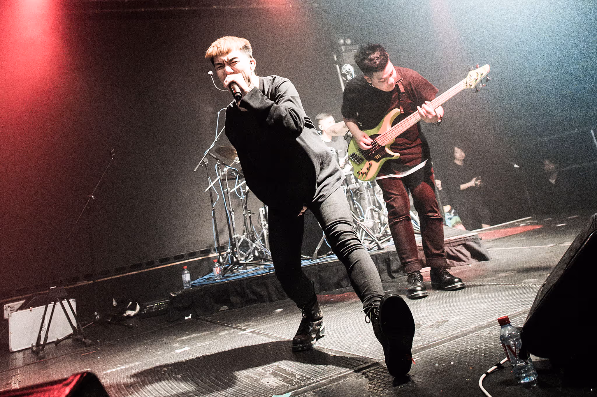 Male vocalist passionately singing into a microphone on stage with a bassist playing behind him under dramatic lighting.