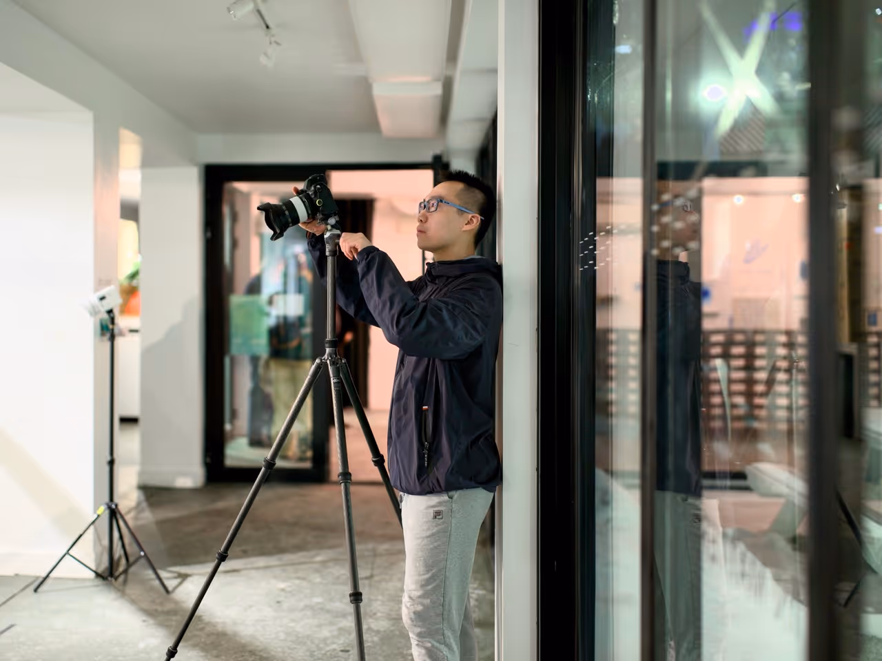 Person adjusting camera on a tripod inside a modern room with glass windows and photography lighting equipment.
